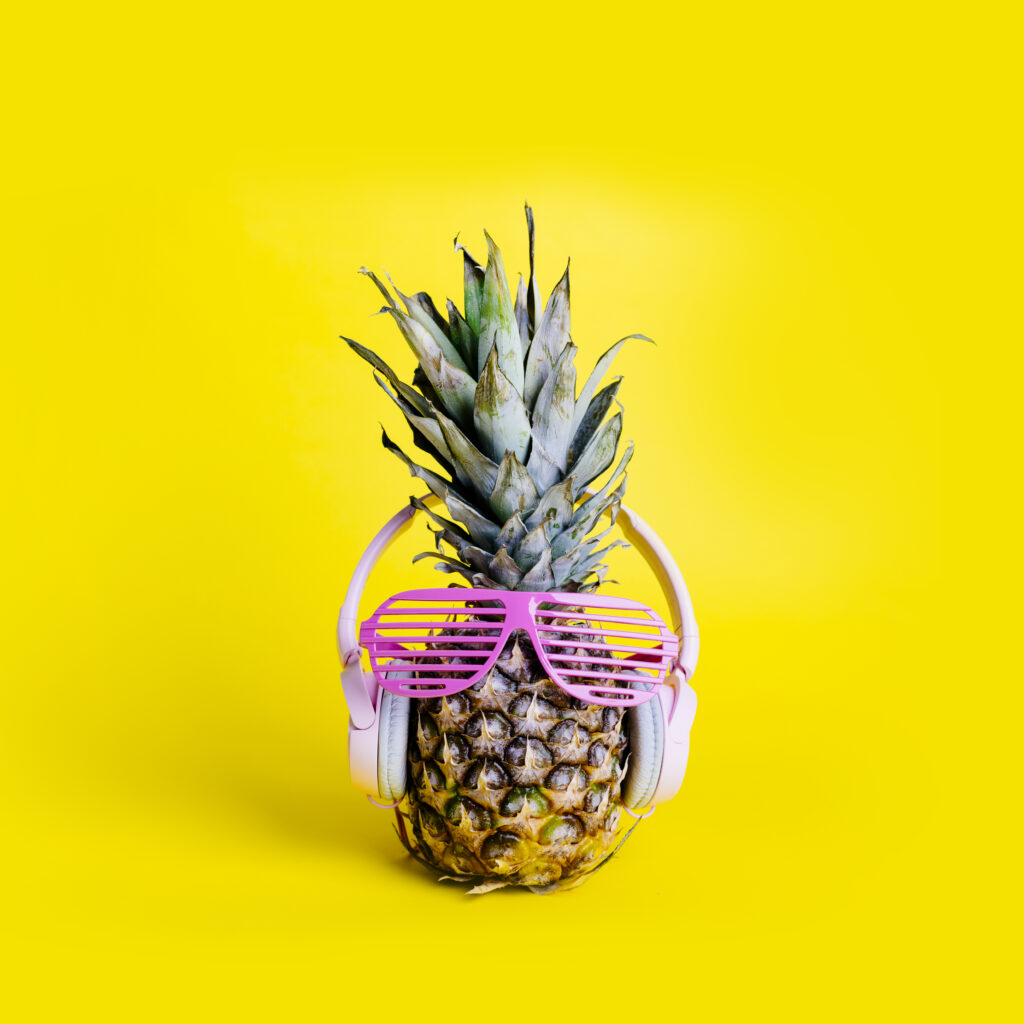 Photo d'un ananas avec une paire de lunette excentrique rose, sur fond jaune.